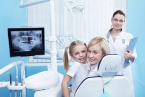 A mom with her daughter sit in a dental chair. A dental hygienist is caring for them. A solid patient recall system keeps the dental hygiene chairs fully booked.
