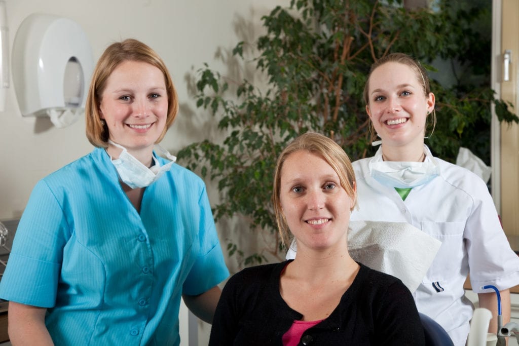 A Dental Front Office Team Member Is Seated At The Dental Front Office. A Dental Assistant And A Dental Hygienist Stand Behind Her. They Have Been Successful In Completing And Implementing Dental Front Desk Management Systems Course