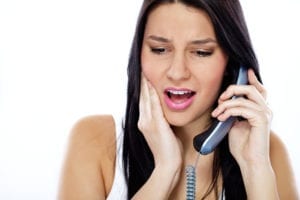 A woman holds the phone to her ear and holds her cheek. She is calling the dentist to schedule a new patient appointment.
