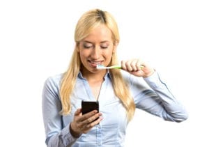 A Female Dental Patient Brushing Her Teeth & Reading Texted Appointment Confirmation. Stronger Appointment Confirmations Reduce The Number of Patients Cancelling Dentist Appointments Last Minute