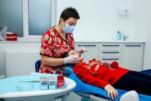 Seasonal Dental Practice Management Is most successful when we embrace the season before us and use the season to care for our patients and the dental team. A female dentist is wearing a Christmas uniform and placing a sealant on the young dental patient's tooth.