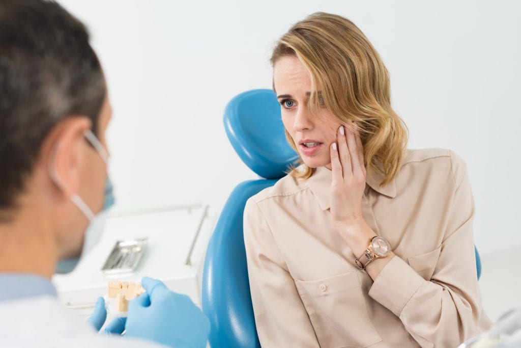 A dentist wearing a mask sits next to a female patient in his dental chair. She holds her mouth. Scheduling dental emergency patients well keeps the dental restorative schedule productive and provides exceptional patient care.
