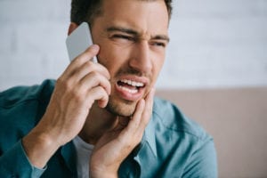 A man holds his cheek and is talking on his cell phone.  He has called the dental office for help.  Scheduling dental emergency patients is usually done over the telephone.  