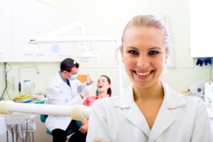 A dentist is working on a female dental patient while a dental assistant looks toward us and smiles.  Scheduling dental emergency patients well grows the dental practice.
