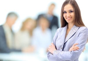 A female professional stands in front of a group of other professionals who are faded out in the background. Learning dental restorative scheduling takes time, education, and experience.