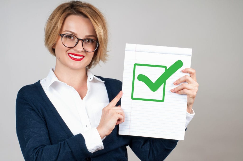 A smiling business woman holds a white paper with a green check mark on it.