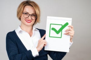 A smiling business woman holds a white paper with a green check mark on it. She has completed a credit balance audit and is able to close her month.