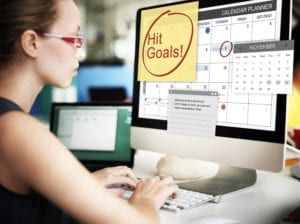 A Dental Front Office Team Member Sits At Her Computer With A Big Note On Her Screen That Says "Hit Goals". She Is Setting Up Her Tracking System To Reach Monthly Production Goals.